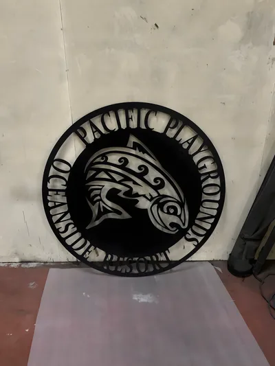 Black circular metal sign reading Pacific Playgrounds Oceanside Resort with a fish design
